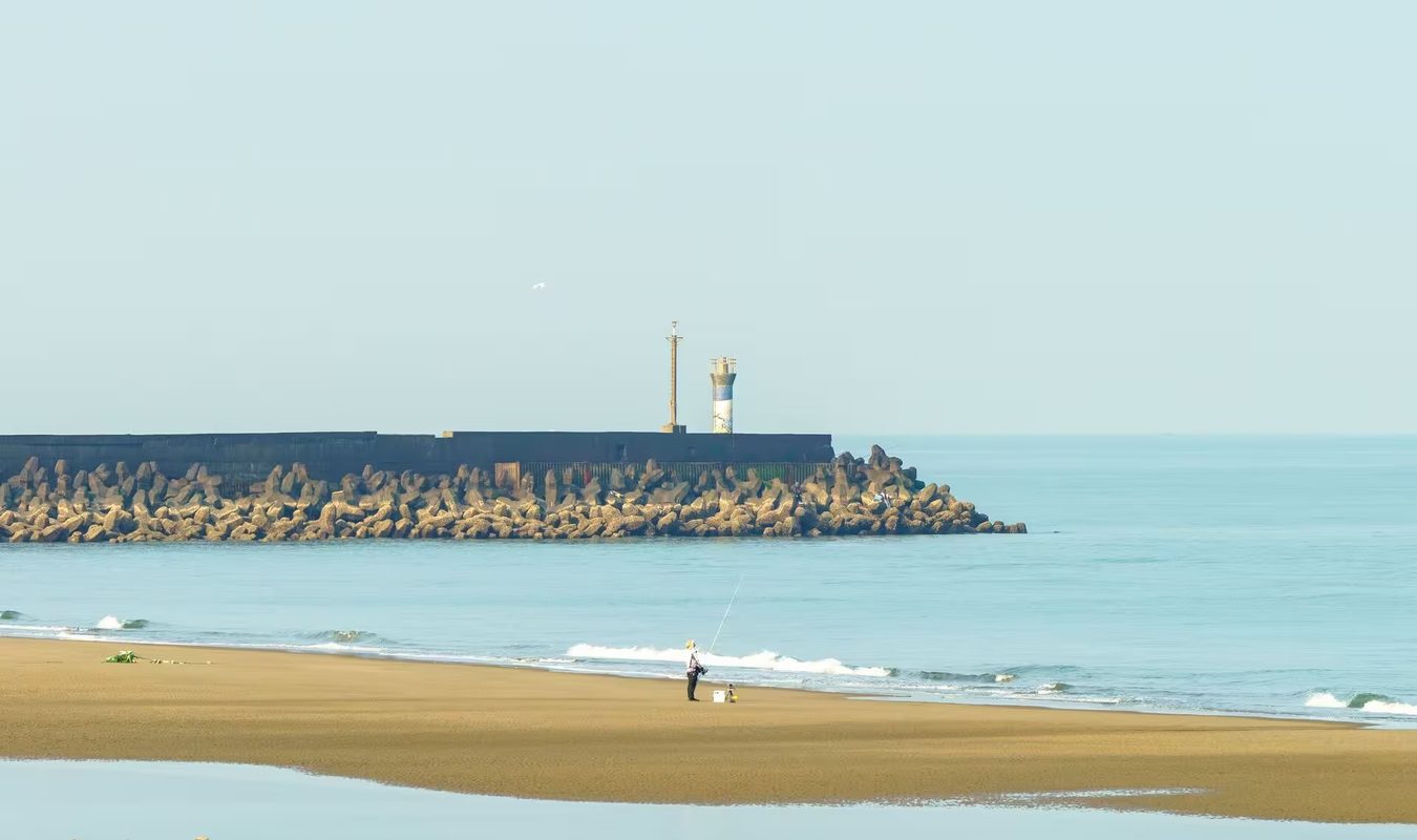 Une plage d'Anglet à marée basse, la digue de blocs de béton et son petit phare au bout, un pêcheur seul sur le sable face aux vagues
