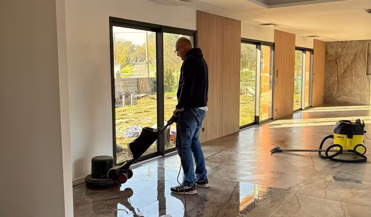 Une pièce aux grandes baies vitrées, juste après les travaux : un intervenant passe la monobrosse sur le carrelage, un aspirateur eau et poussières au fond.