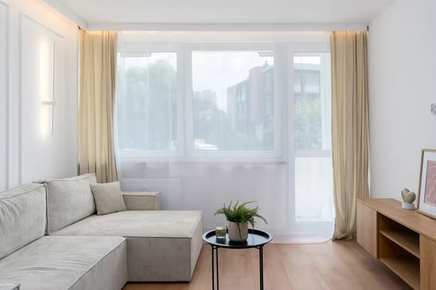 Un séjour lumineux, une baie vitrée voilée de blanc derrière un canapé d'angle gris