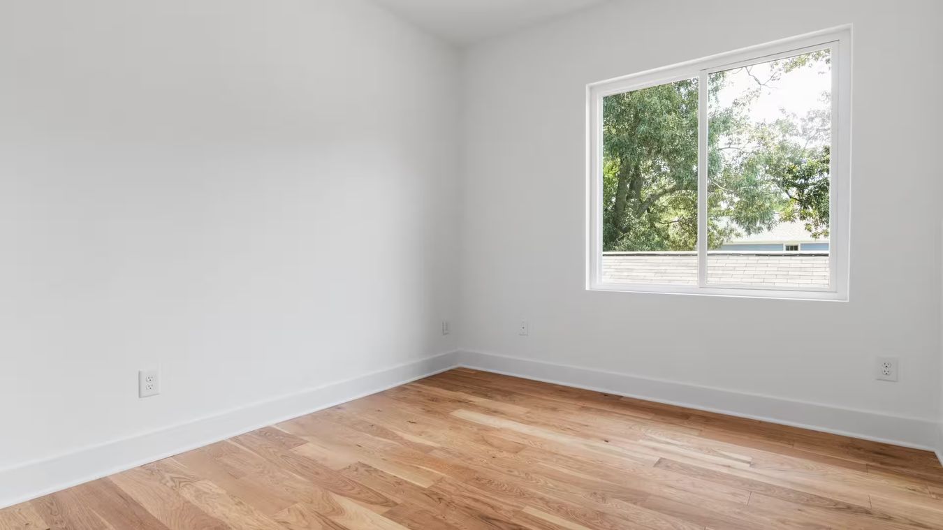 Une pièce vide aux murs blancs et au parquet clair, une grande fenêtre ouverte sur les arbres