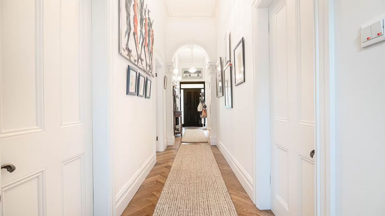 Un long tapis de couloir sur un parquet à chevrons, entre des murs blancs ornés de cadres