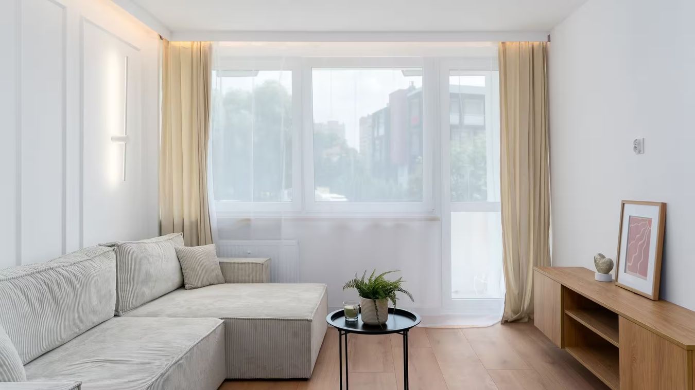 Un séjour lumineux, une baie vitrée voilée de blanc derrière un canapé d'angle gris