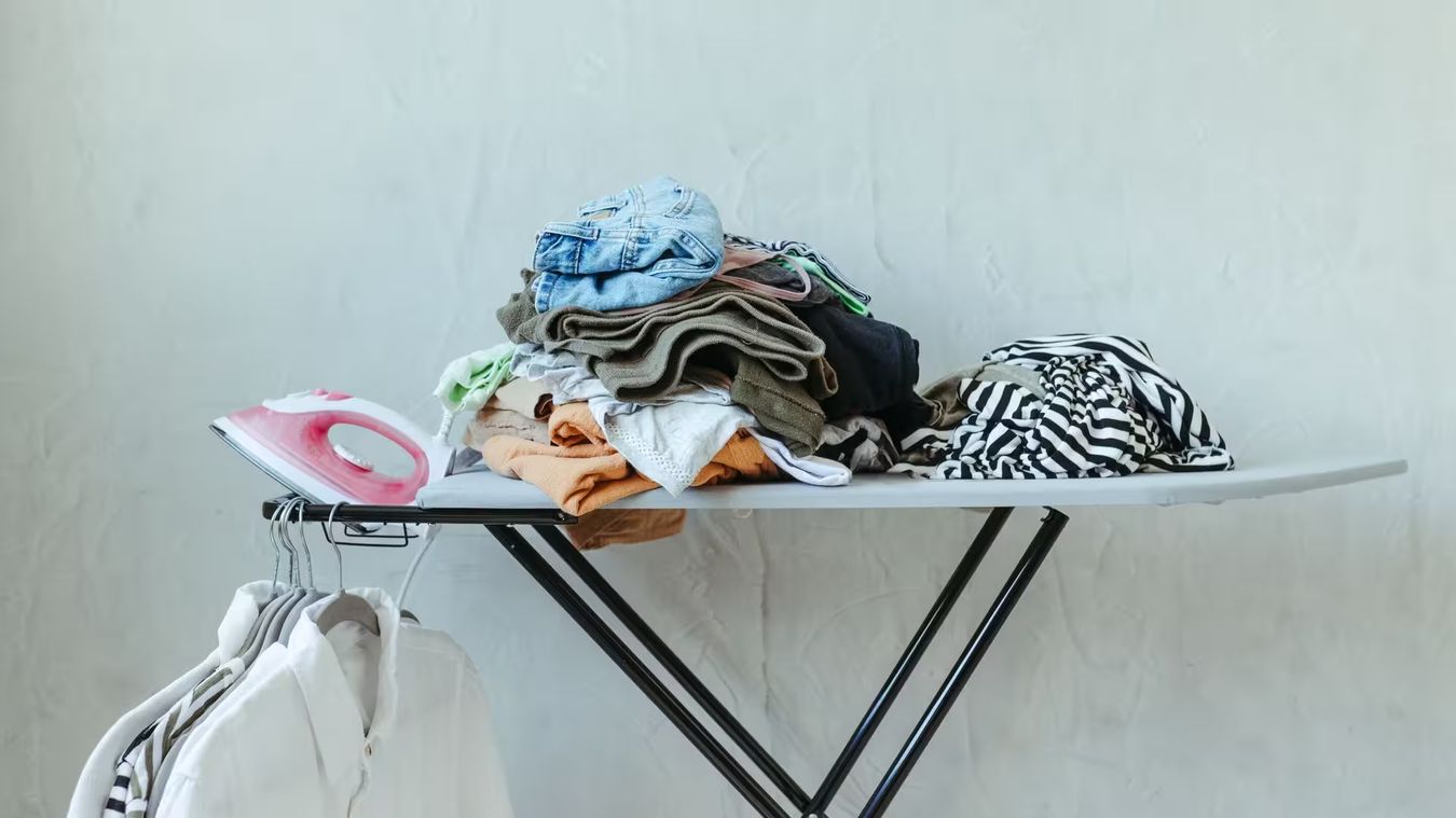 Une pile de linge et un fer posés sur une planche à repasser, des chemises suspendues à côté