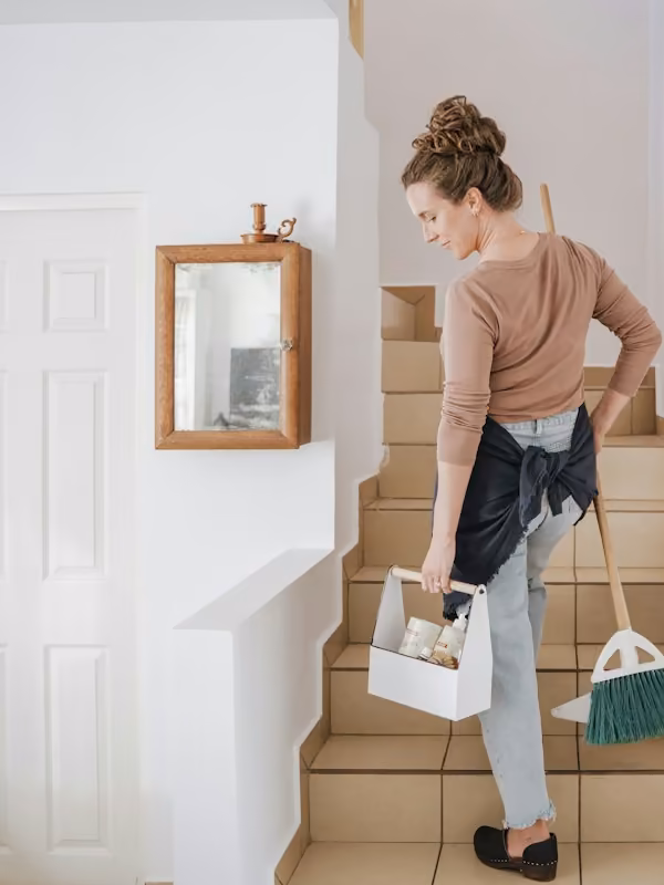 Une femme monte un escalier, caisse de produits et balai à la main